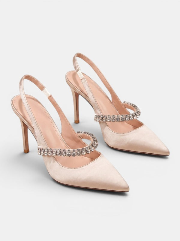 A pair of pointed-toe slingback high heels in pale blush pink satin, featuring a decorative band of clear rhinestones across the instep and around the ankle straps, set against a light gray studio background.